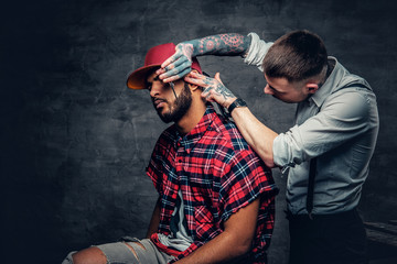 Naklejka premium Tattooed barber cutting the beard of a Black hipster male.
