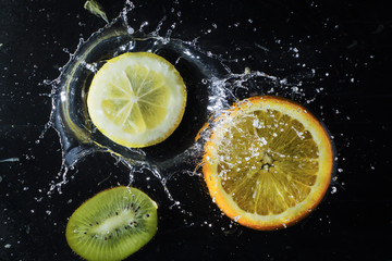 Citrus in water on a black background. Splashing water droplets. Kiwi, lemon and orange