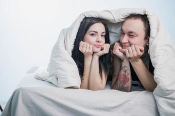 brunette girl and the guy in the tattoo lay under a white blanket wrapped with a smile looking at camera on white background copyspace