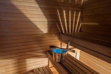 Fototapeta na wymiar Wellness and spa conception. Sauna bath bucket with blue water, scoop, striped light, wooden walls and timber bench