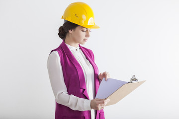 Close up portrait of a serious woman architect wearing a pink vest and a yellow safety helmet and holding a clipboard with construction documents.