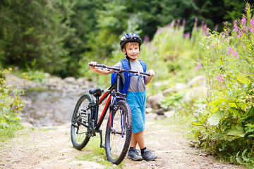 Fototapeta na wymiar Small smiling boy in helmet standing with bicycle on mountain trail