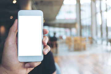 Mockup image of hand holding white mobile phone with blank white screen in modern cafe