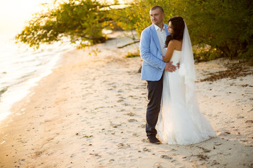 Portrait of Couple of lovers newlyweds on the seafront. Brunette bride in wedding dress with a beautiful hairstyle, veil and makeup and her husband