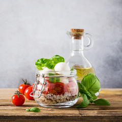 Delicious caprese salad with quinoa, ripe cherry tomatoes and mini mozzarella cheese with fresh basil leaves in glass jar and olive oil. Italian healthy food concept with copy space.