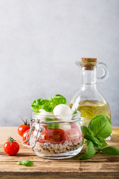 Delicious caprese salad with quinoa, ripe cherry tomatoes and mini mozzarella cheese with fresh basil leaves in glass jar and olive oil. Italian healthy food concept with copy space.