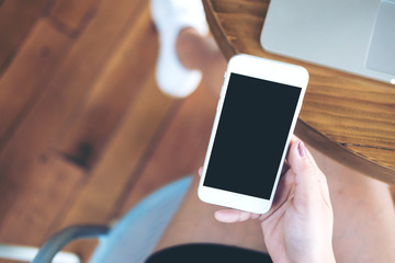 Mockup image of hand holding white mobile phone with blank black screen on thigh and silver colour laptop on table with wooden floor background