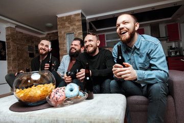 A company of four men having fun and laughing sitting on the couch at home with beer and chips, two of them with joysticks in hand playing computer video game.