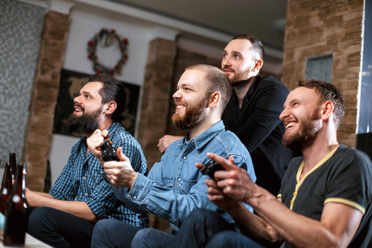 Men with a beard sitting on the couch at home with beer and chips with joysticks in hand playing computer video games. The concept of friendship, technology and weekend