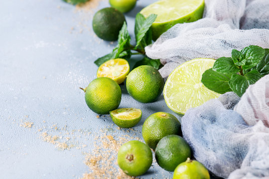 Ingredients for mojito cocktail, whole, sliced lime and mini limes, mint leaves, brown crystal sugar over gray stone texture background with gauze textile. Copy space. Close up