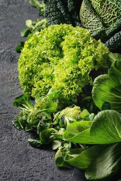 Variety of raw green vegetables salads, lettuce, bok choy, corn, broccoli, savoy cabbage as frame over black stone texture background. Space for text