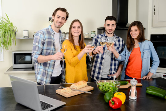 group of friends, four people man and woman at home cooking, drinking wine and preparing food meal together in the kitchen