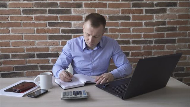 Man working on the laptop and writing something in the notebook, brick wall background