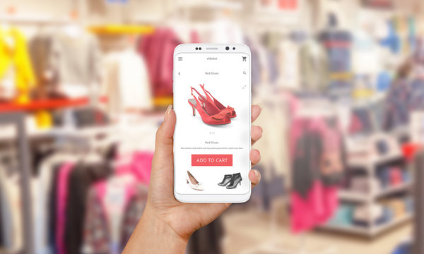 Online shopping with mobile phone. PHone in woman hand. Shop market online app on screen. Clothes and footwear shop in background.