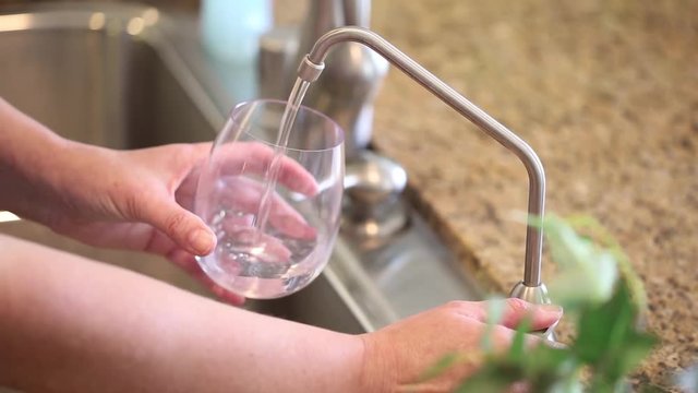 Woman Pouring Fresh Purified Reverse Osmosis Drinking Water Into Clear Glass at Kitchen Sink Faucet