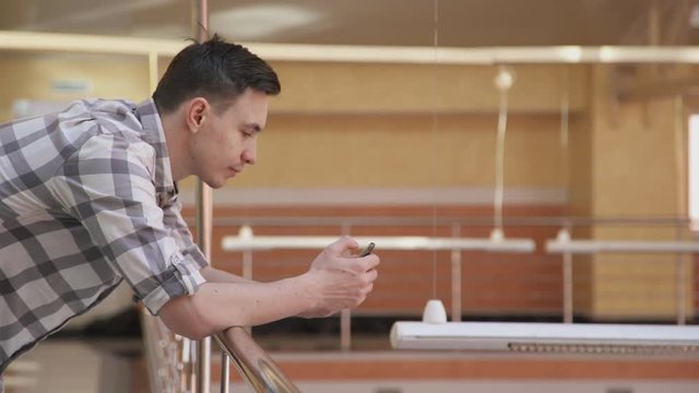 man holding two phones work looking for information in office. Male with brown hair and gray plaid white shirt standing on second floor in hall, leaning against railing, looking at smart phone, takes