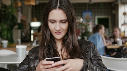 Stylish young woman uses a smartphone sends sms sitting at a table in modern cafe, smiling. 4K