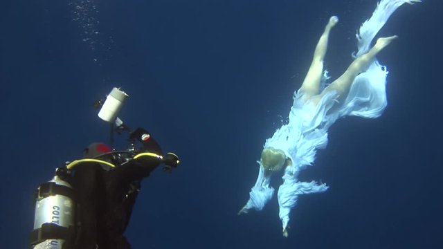 Underwater model free diver in costume angel swims in clean water in Red Sea. Filming a movie. Young girl smiling at camera. Extreme sport in marine landscape, coral reefs, ocean inhabitants.