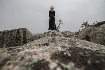Artistic photo of a blond girl in a transparent black dress walking on the rocks in the field a cloudy day. The aesthetic is sinister and a little dark. 