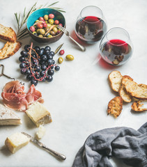 Wine and snack set. Variety of cheese, olives in ceramic bowl, prosciutto, roasted baguette slices, black grapes and glasses of red wine over grey marble background, selective focus, copy space