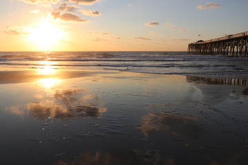 Naklejka premium Atlantic ocean sunrise background. Sunrise over the ocean. Atlantic ocean landscape with a sun and clouds reflected in shallow water and wooden pier in Myrtle Beach area, South Carolina, USA.