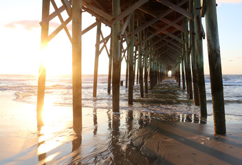 Fototapeta premium Atlantic ocean sunrise background. Sunrise over the ocean. Atlantic ocean landscape with a sun and clouds reflected in shallow water and wooden pier in Myrtle Beach area, South Carolina, USA.