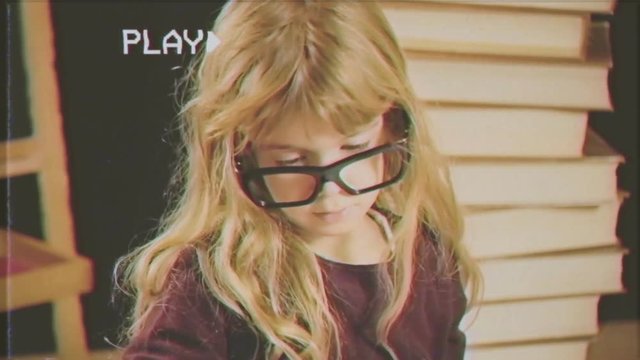 Fake VHS tape: a pretty little blonde girl with nerdy glasses is busy reading & studying. A big pile of books and a blackboard are behind her.
