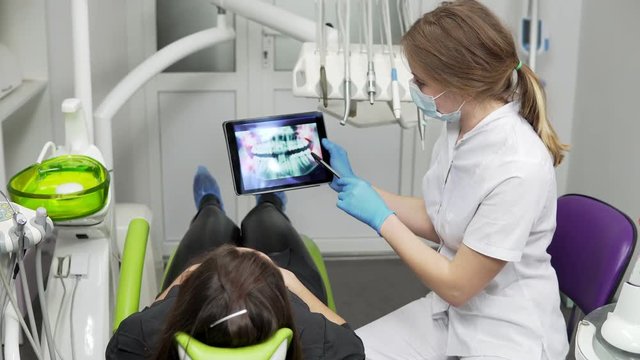 Female doctor dentist is showing x-ray teeth on tablet to young female patient. Dentist is wearing lab coat
