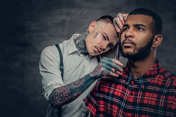 Tattooed barber cuts the beard to a Black bearded male.