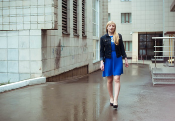 Blond woman in blue dress and denim jacket walking down the city street against a background of urban architecture