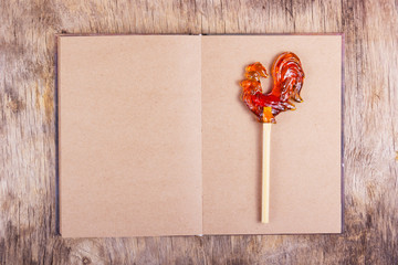 Open book with blank pages and a rooster on a stick. Lollipop on a stick in the form of a rooster on an empty sheet. Copy space.