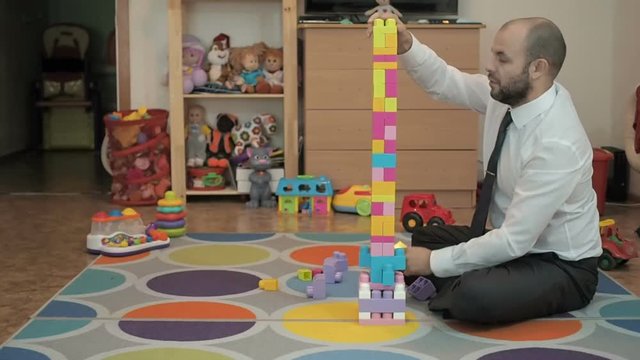 Adult male businessman collects children's building-block and enjoys sitting on the floor in the nursery.