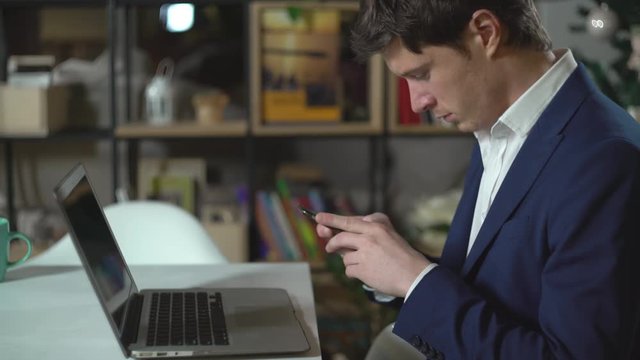 Handsome man sitting in room with bookshelves on the desk laptop. Elegant fashionable man wearing in casual blue suit texting message on smartphone.