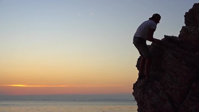 Young man climbs on the rock against colourful sunset background