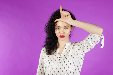 young curly brunette woman giving loser sign on forehead on a pink background