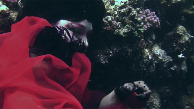 Young girl model free diver underwater in red costume of pirate in Red Sea. Filming a movie. Actress smiling at camera. Extreme sport in marine landscape, coral reefs, ocean inhabitants.