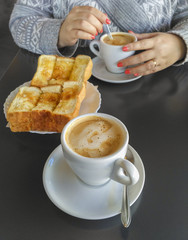 Hands of woman holding cup of coffee with milk and toast with butter