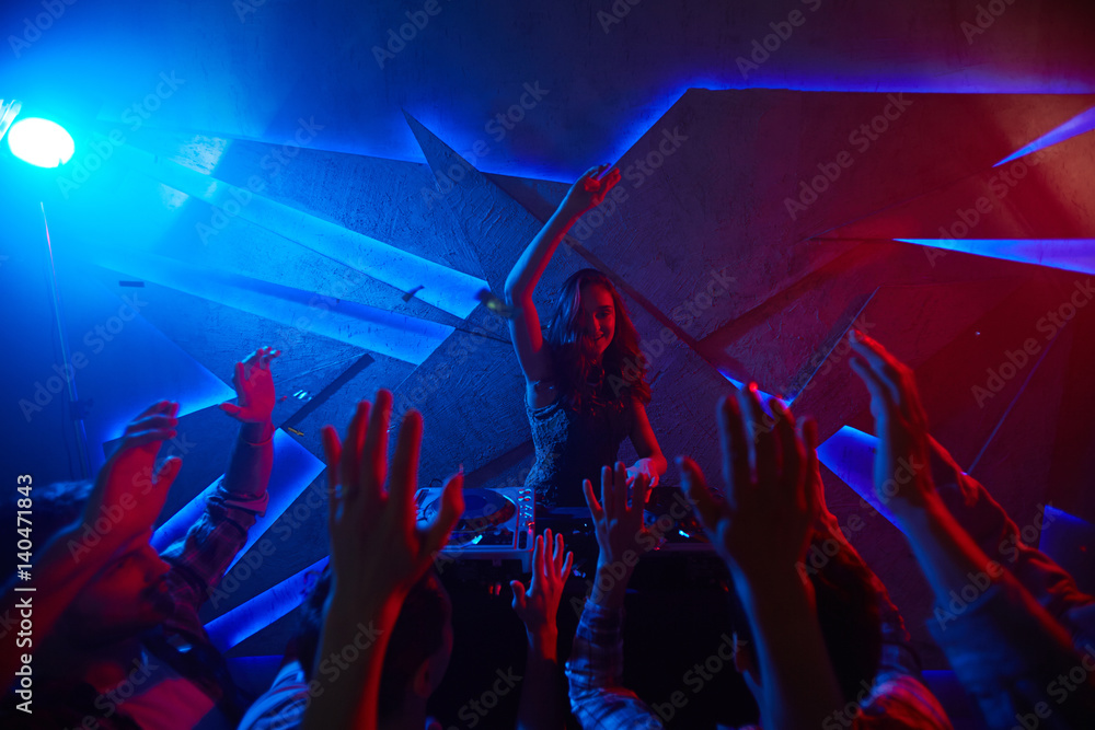 Poster Girl raising hand while standing by turntables in front of dancers