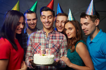 Happy young friends looking at burning candles on tasty birthday cake