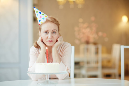 Portrait Of Sad  Lonely Mature Woman Sitting Alone At Birthday Table With Cake , Wearing Party Hat And Looking At Camera