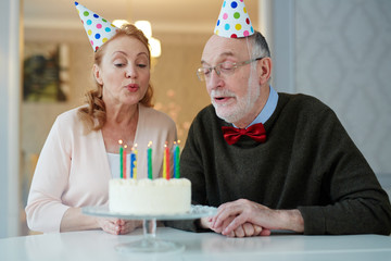 Portrait of loving senior couple celebrating birthday together sitting at table with cake and wearing party hats blowing out candles and making wishes