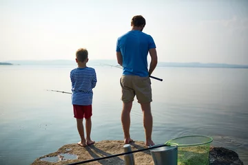 Foto op Plexiglas Back view portrait of muscular well-built man adjusting fishing rod while standing on lake with his little son, sharing hobby on sunny summer day © pressmaster