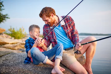 Papier Peint photo autocollant Pêcher Portrait d& 39 un beau père ludique chatouillant son fils assis sur un rocher au bord du lac tout en profitant de la pêche ensemble et en riant dans les rayons du soleil couchant