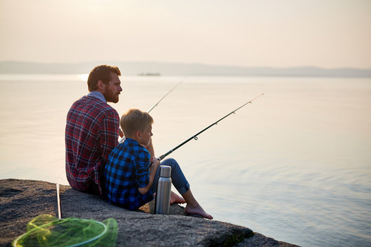 Back view portrait of adult man and teenage boy sitting together on rocks fishing with rods in calm waters of blue lake at dusk, both wearing checkered shirts