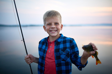 Portrait of blond happy boy smiling  looking at camera holding fishing rod and single perch against calm blue lake background at dusk