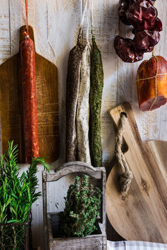 Charcuterie, variety of sausages hanging on hook, wood cutting board, string with dry peppers, fresh garden herbs
