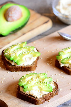 Hummus avocado sandwiches on a wooden board, avocado half. Sandwiches cooked with rye bread, fresh avocado slices, hummus and roasted sesame seeds. Vertical photo