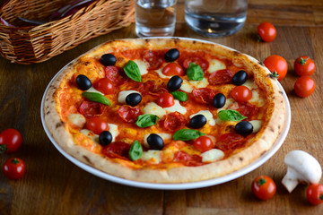 Tasty hot pizza with pepperoni, mozzarella, cherry tomatoes and basil, served on a wooden table for a dinner in restaurant. Pizza from wooden oven. Italy food concept.