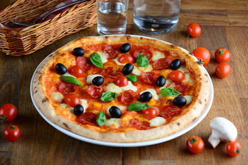 Tasty hot pizza with pepperoni, mozzarella, cherry tomatoes and basil, served on a wooden table for a dinner in restaurant. Pizza from wooden oven. Italy food concept.