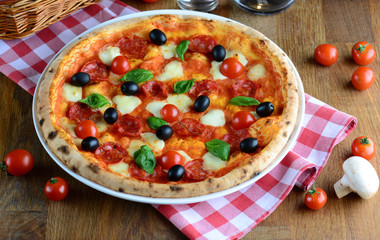 Hot tasty pizza with pepperoni, mozzarella, cherry tomatoes and basil, served on a wooden table for a dinner in italian restaurant. Pizza from wooden oven. Italy food concept.
